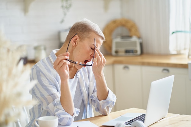 Mujer de mediana edad que se masajea la nariz y se siente somnolienta mientras trabaja en la computadora portátil en línea desde casa