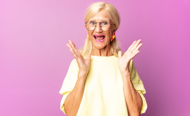 Mujer de mediana edad que parece feliz y emocionada, sorprendida con una sorpresa inesperada con ambas manos abiertas junto a la cara