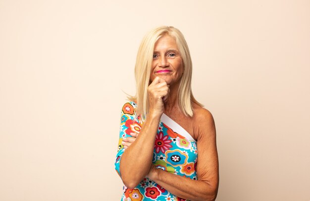 Mujer de mediana edad que parece feliz y sonriente con la mano en la barbilla, preguntándose o haciendo una pregunta, comparando opciones