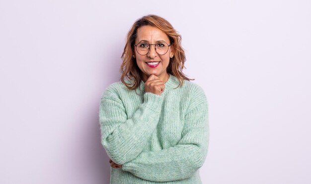 Mujer de mediana edad que parece feliz y sonriente con la mano en la barbilla, preguntándose o haciendo una pregunta, comparando opciones