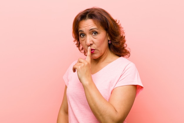 Mujer de mediana edad que pide silencio y silencio, gesticulando con el dedo frente a la boca, diciendo shh o guardando un secreto contra la pared rosa