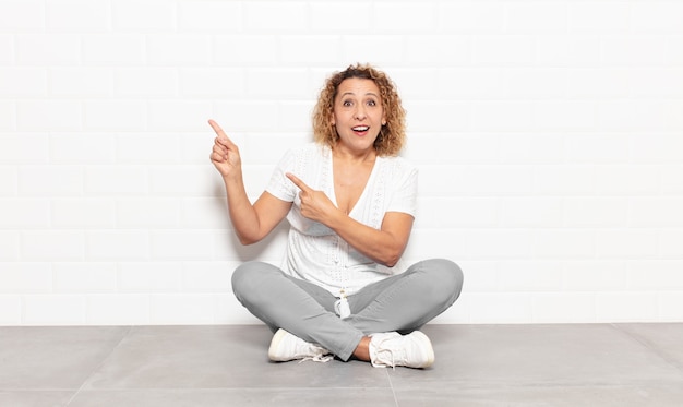 Mujer de mediana edad que se siente alegre y sorprendida, sonriendo con una expresión de sorpresa y apuntando hacia un lado