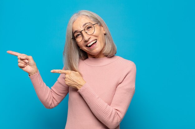 Mujer de mediana edad que se siente alegre y sorprendida, sonriendo con una expresión de sorpresa y apuntando hacia un lado