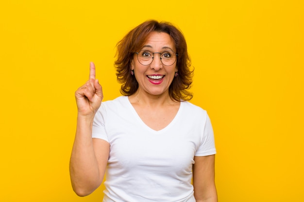 Mujer de mediana edad que se siente como un genio feliz y emocionado después de darse cuenta de una idea, levantando alegremente el dedo, ¡eureka! contra la pared amarilla