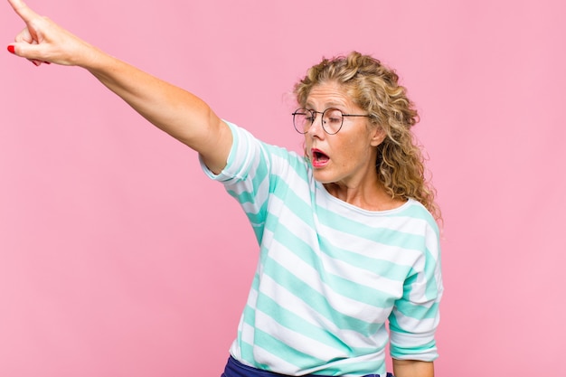 Mujer de mediana edad que se siente conmocionada y sorprendida, señalando y mirando hacia arriba con asombro con una mirada asombrada y con la boca abierta.