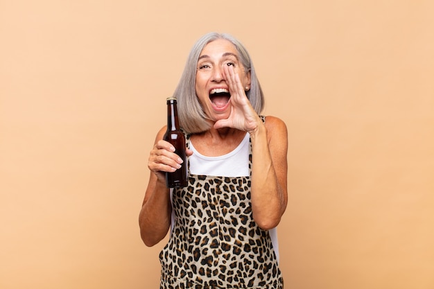 Mujer de mediana edad que se siente feliz, emocionada y positiva, dando un gran grito con las manos al lado de la boca, gritando con una cerveza