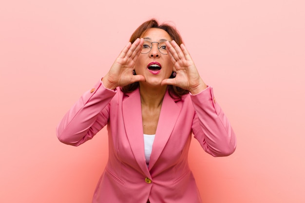 Mujer de mediana edad que se siente feliz, emocionada y positiva, dando un gran grito con las manos al lado de la boca, gritando pared rosa