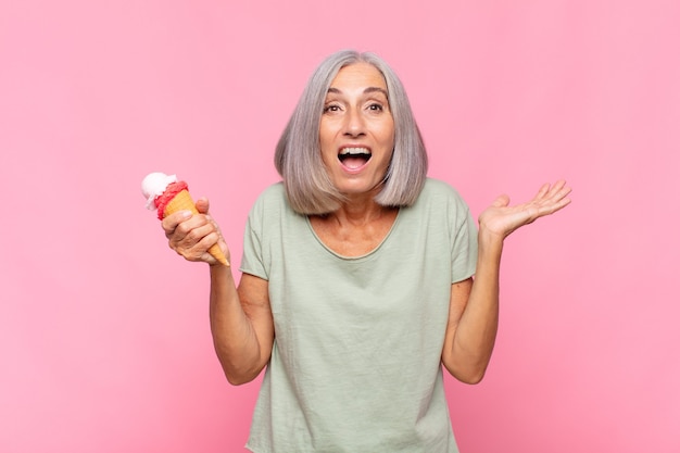 Mujer de mediana edad que se siente feliz, emocionada, sorprendida o conmocionada, sonriendo y asombrada por algo increíble tomando un helado