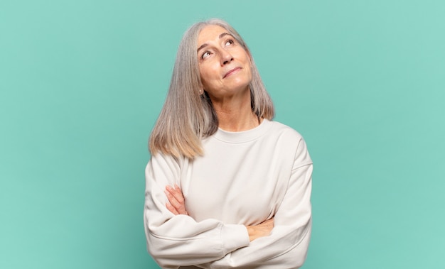 Mujer de mediana edad que se siente feliz, orgullosa y esperanzada, preguntándose o pensando, mirando hacia arriba para copiar el espacio con los brazos cruzados