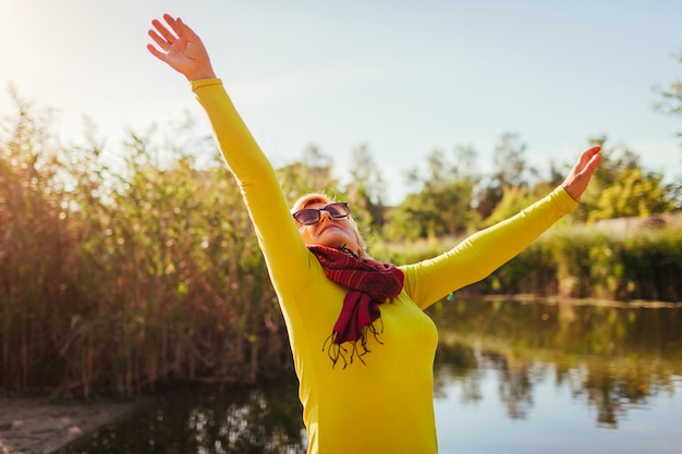 Mujer de mediana edad que se siente libre y feliz en la orilla del río el día de otoño. Señora mayor levantando las manos. Armonía y equilibrio con la naturaleza