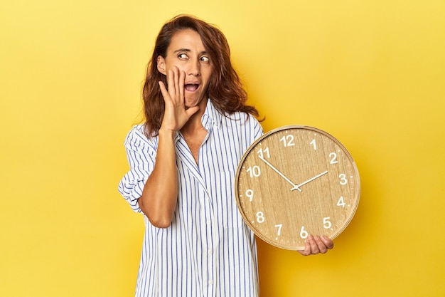 Una mujer de mediana edad que sostiene un reloj de pared sobre un fondo amarillo está diciendo una noticia secreta sobre el frenado en caliente