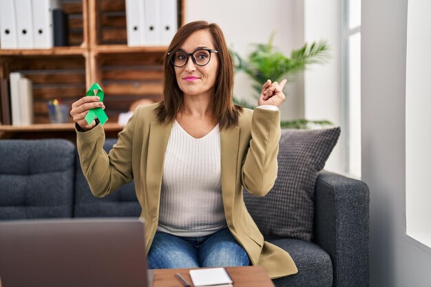 Mujer de mediana edad que trabaja sosteniendo la cinta verde de apoyo sonriendo feliz señalando con la mano y el dedo hacia un lado