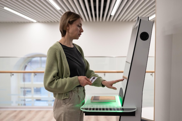 Mujer de mediana edad registrando un libro en la biblioteca usando una terminal de autoservicio