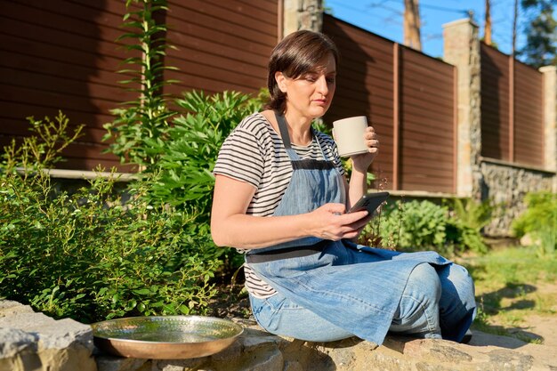 Mujer de mediana edad relajándose en el jardín con un teléfono inteligente y una taza en las manos