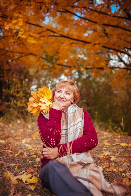 Mujer de mediana edad rodeada de árboles amarillos se relaja en el bosque de otoño. Señora disfrutando de la naturaleza con ramo de hojas