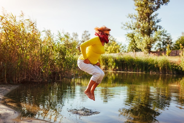 Mujer de mediana edad saltando en la orilla del río el día de otoño. Señora mayor feliz divirtiéndose caminando en el bosque. Sentirse enérgico y libre