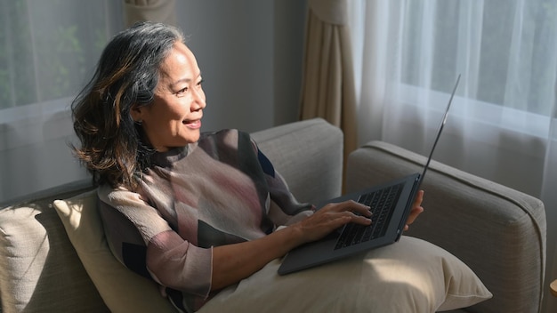 Mujer de mediana edad satisfecha que usa una computadora portátil en un cómodo sofá en casa Concepto de tecnología de jubilación