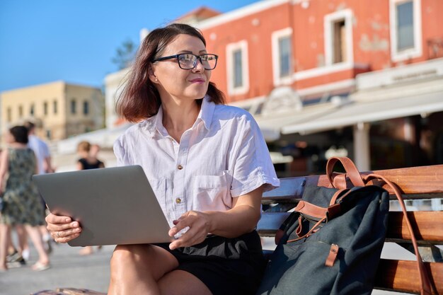 Mujer de mediana edad sentada en un banco en la ciudad usando una computadora portátil