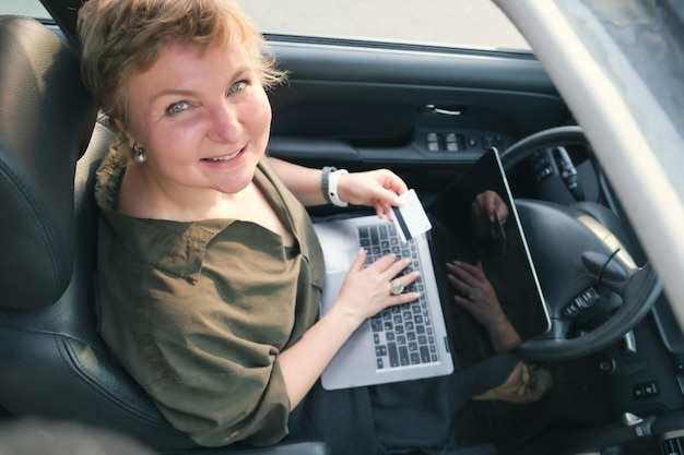 Mujer de mediana edad se sienta al volante de un automóvil con una computadora portátil en su regazo