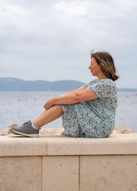 Una mujer de mediana edad se sienta en la orilla del mar y mira a la distancia el mar y las montañas en el horizonte