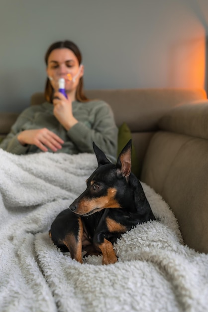 Una mujer de mediana edad se sienta en un sofá y hace una inhalación a través de un nebulizador junto a un perro.