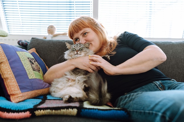 Una mujer de mediana edad se sienta en el sofá junto a la ventana con un gato