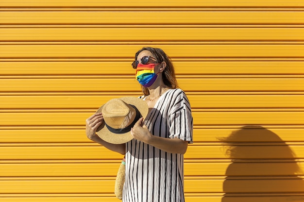 Mujer de mediana edad con sombrero, gafas de sol y una máscara protectora con los colores del arco iris. LGTB sobre fondo amarillo. Concepto Covid-19