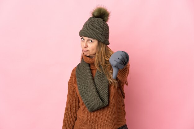 Foto mujer de mediana edad con sombrero de invierno aislado sobre fondo rosa mostrando el pulgar hacia abajo con expresión negativa