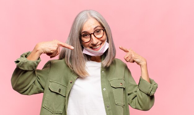 Mujer de mediana edad sonriendo con confianza apuntando a su propia sonrisa amplia, actitud positiva, relajada y satisfecha