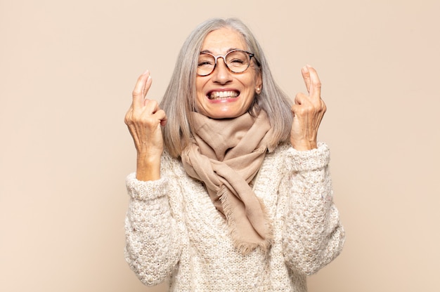 Mujer de mediana edad sonriendo y cruzando ambos dedos con ansiedad, sintiéndose preocupada y deseando o esperando buena suerte