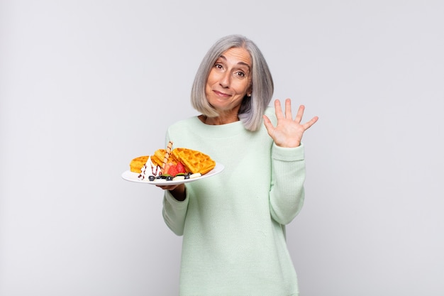 Mujer de mediana edad sonriendo feliz y alegremente, saludando con la mano, dándole la bienvenida y saludándolo, o despidiéndose