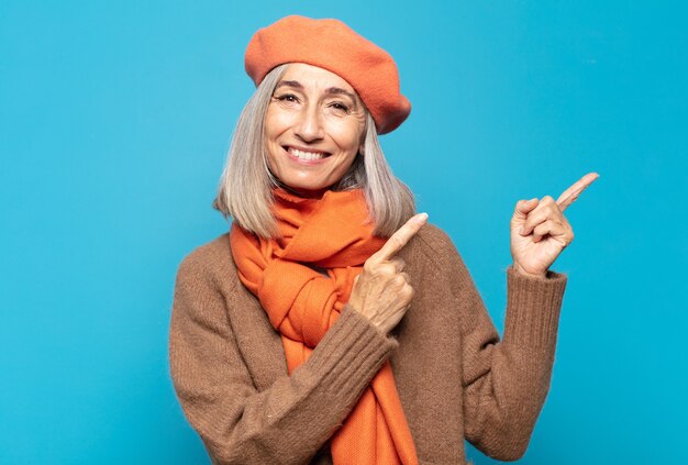 Mujer de mediana edad sonriendo felizmente y apuntando hacia un lado y hacia arriba con ambas manos mostrando el objeto en el espacio de la copia