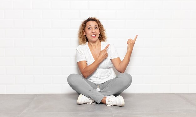 Mujer de mediana edad sonriendo felizmente y apuntando hacia un lado y hacia arriba con ambas manos mostrando el objeto en el espacio de la copia