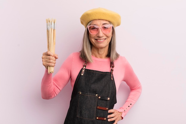 Mujer de mediana edad sonriendo felizmente con una mano en la cadera y un concepto de pintor de artista confiado