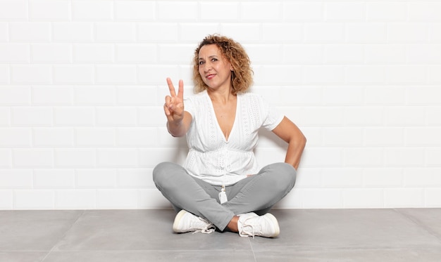 Mujer de mediana edad sonriendo y luciendo feliz, despreocupada y positiva, gesticulando victoria o paz con una mano