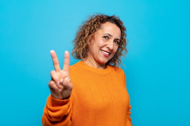 Mujer de mediana edad sonriendo y luciendo feliz, despreocupada y positiva, gesticulando victoria o paz con una mano