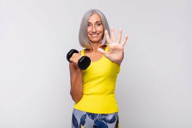 Mujer de mediana edad sonriendo y mirando amistosamente, mostrando el número cinco o quinto con la mano hacia adelante, contando hacia atrás. concepto de fitness