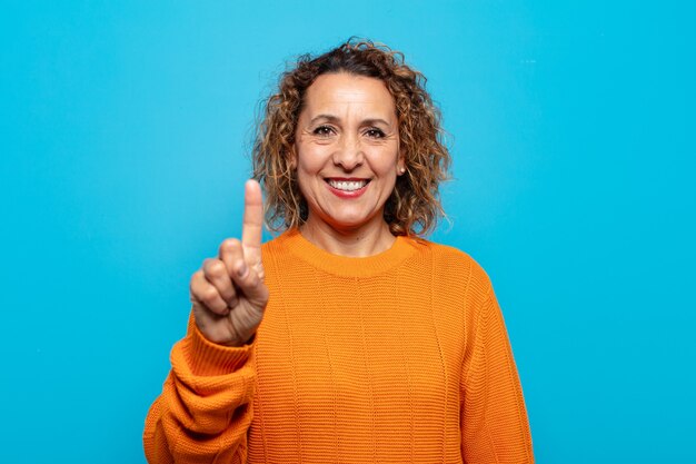 Foto mujer de mediana edad sonriendo y mirando amistosamente, mostrando el número uno o el primero con la mano hacia adelante, contando hacia atrás