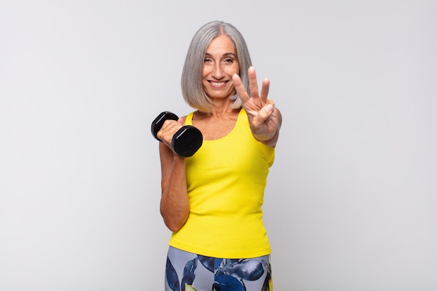 Mujer de mediana edad sonriendo y mirando amistosamente, mostrando el número tres o tercero con la mano hacia adelante, contando hacia atrás. concepto de fitness