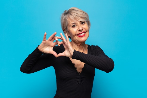 Mujer de mediana edad sonriendo y sintiéndose feliz, linda, romántica y enamorada, haciendo forma de corazón con ambas manos