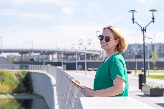 Una mujer de mediana edad sonriente con gafas de sol con un cuaderno o una tableta en las manos en el terraplén de la ciudad Descanso del trabajo