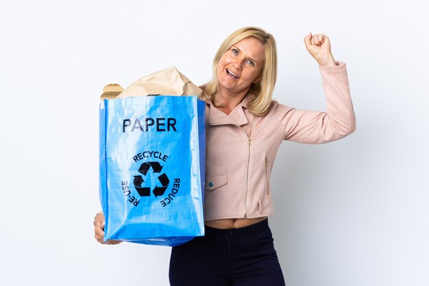 Mujer de mediana edad sosteniendo una bolsa de reciclaje llena de papel para reciclar aislado en la pared blanca celebrando una victoria