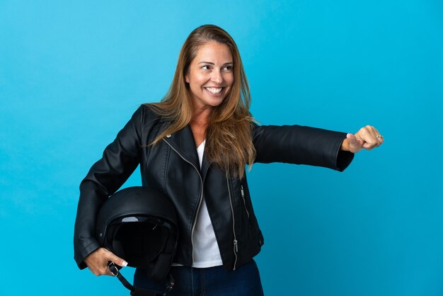 Mujer de mediana edad sosteniendo un casco de motocicleta aislado en la pared azul dando un pulgar hacia arriba gesto