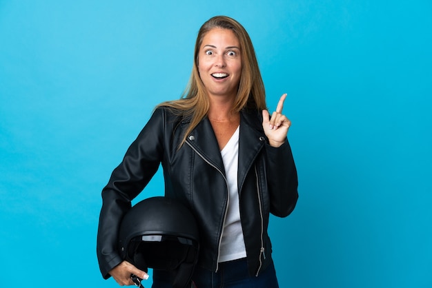 Mujer de mediana edad sosteniendo un casco de motocicleta aislado en la pared azul con la intención de darse cuenta de la solución mientras levanta un dedo