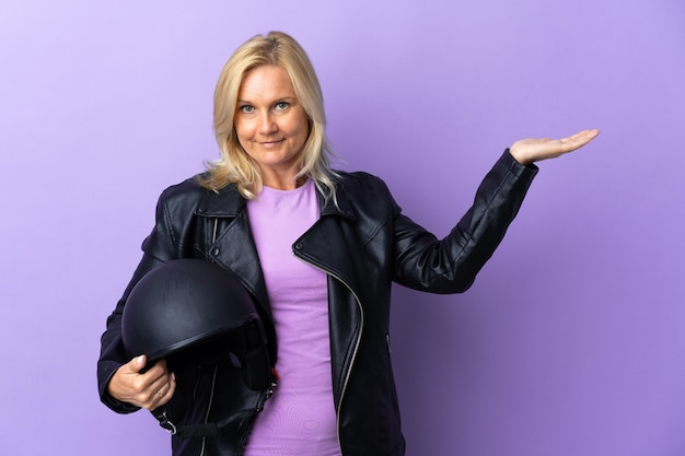 Mujer de mediana edad sosteniendo un casco de motocicleta aislado en la pared púrpura extendiendo las manos hacia un lado para invitar a venir