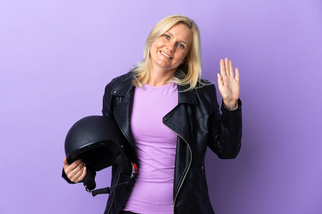 Mujer de mediana edad sosteniendo un casco de motocicleta aislado en la pared púrpura saludando con la mano con expresión feliz