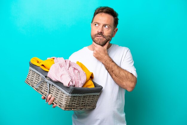 Mujer de mediana edad sosteniendo una cesta de ropa aislada de fondo azul con dudas