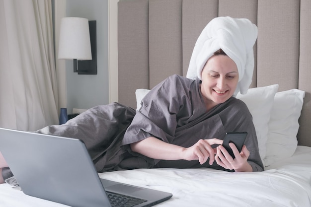 Mujer de mediana edad con una toalla en la cabeza yace en la cama sonriendo y mirando la pantalla del teléfono inteligente