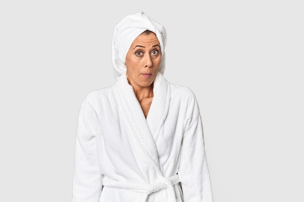 Mujer de mediana edad con toalla después de la ducha en el estudio se encoge de hombros y los ojos abiertos confundidos