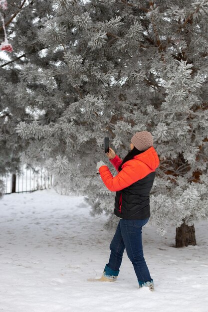 Mujer de mediana edad tomando fotos de árboles con el teléfono parado en un suelo lleno de nieve con árboles y cerca de hierro en el fondo Divirtiéndose afuera en invierno